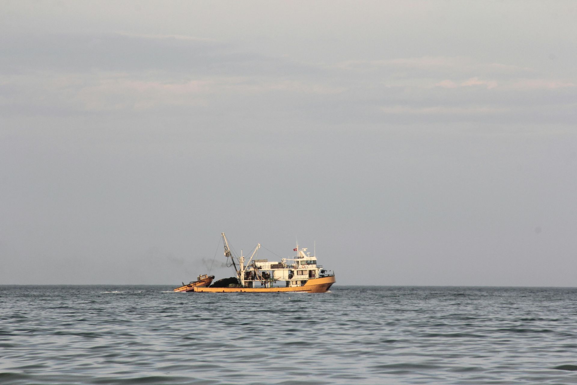 Fishing boat on calm water under a cloudy sky. The boat is light-colored and has various equipment visible.