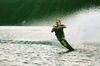 Man water skis on a lake, creating a large spray. He is wearing a dark shirt and shorts. Trees line the background.