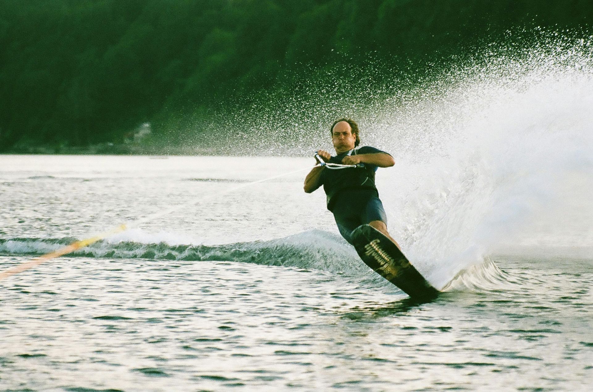 Man water skis on a lake, creating a large spray. He is wearing a dark shirt and shorts. Trees line the background.