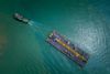 Overhead view: a tugboat tows a large barge across turquoise water. The barge holds equipment and is black with yellow and orange details.