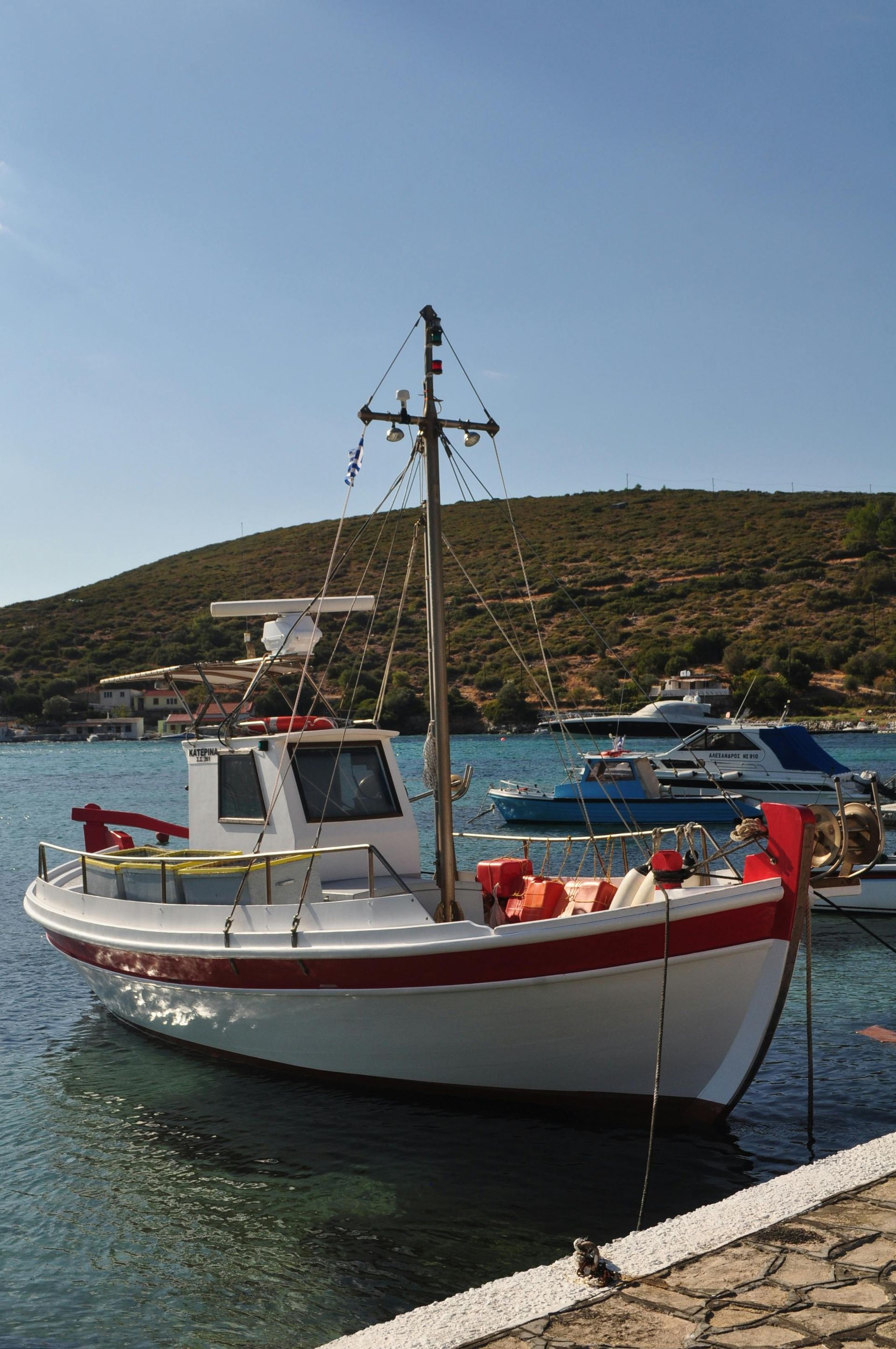 White-and-red fishing boat moored in a harbor with blue water and a hillside in the background