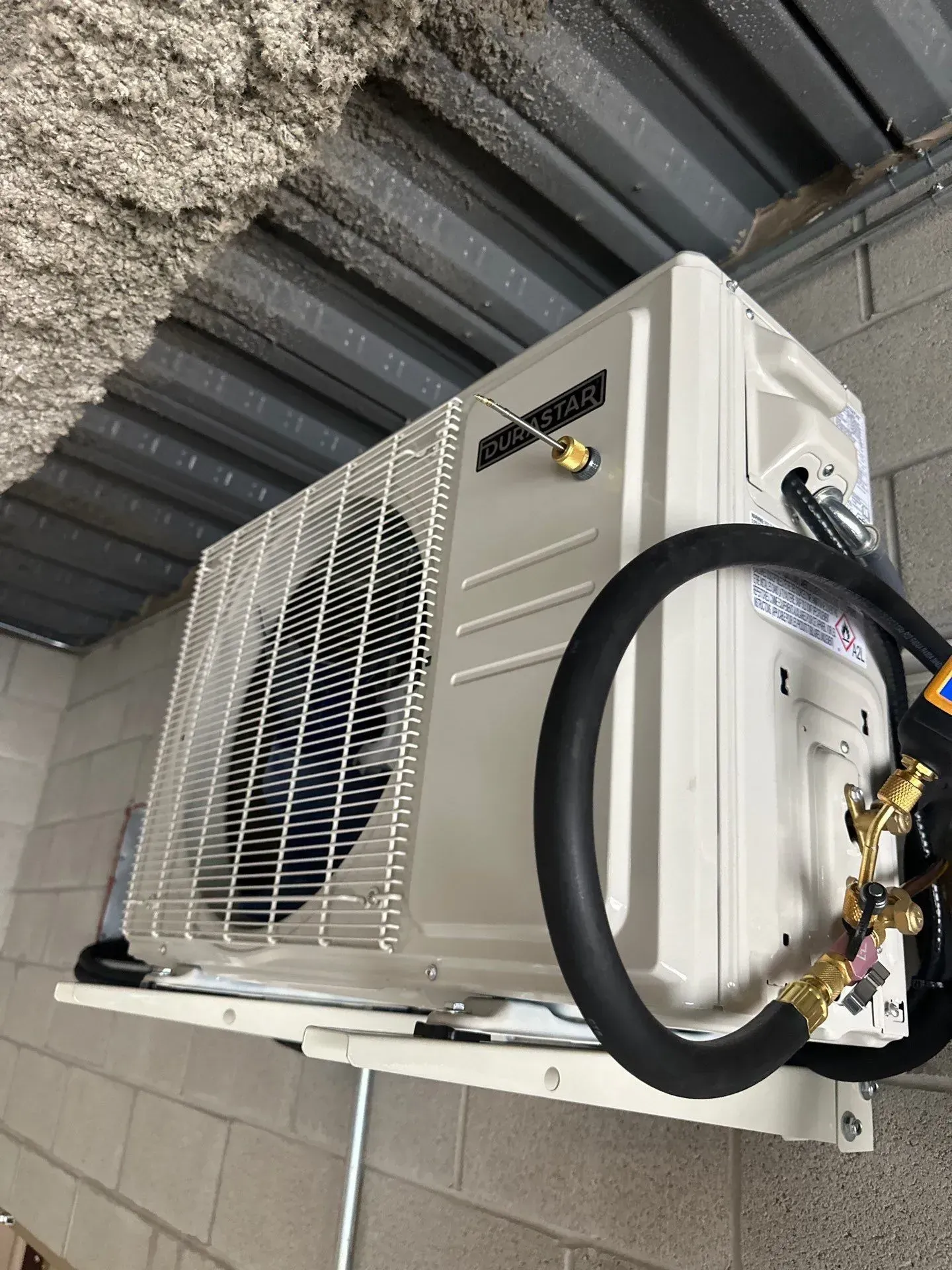 Air conditioning unit mounted on a brick wall, metal ceiling above. Black hose connects to the unit.