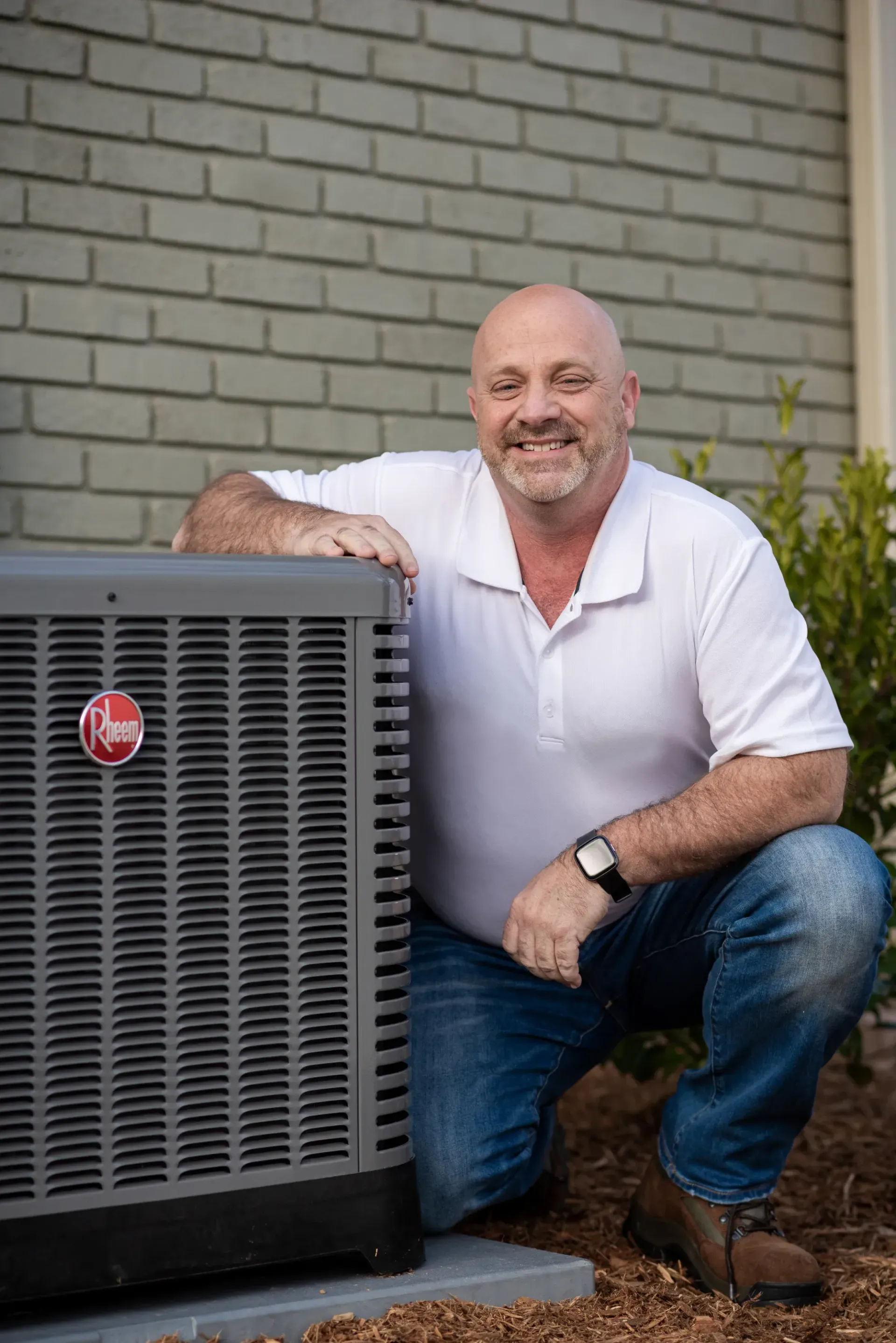 Man in white shirt and jeans, smiling, kneeling next to an air conditioning unit.