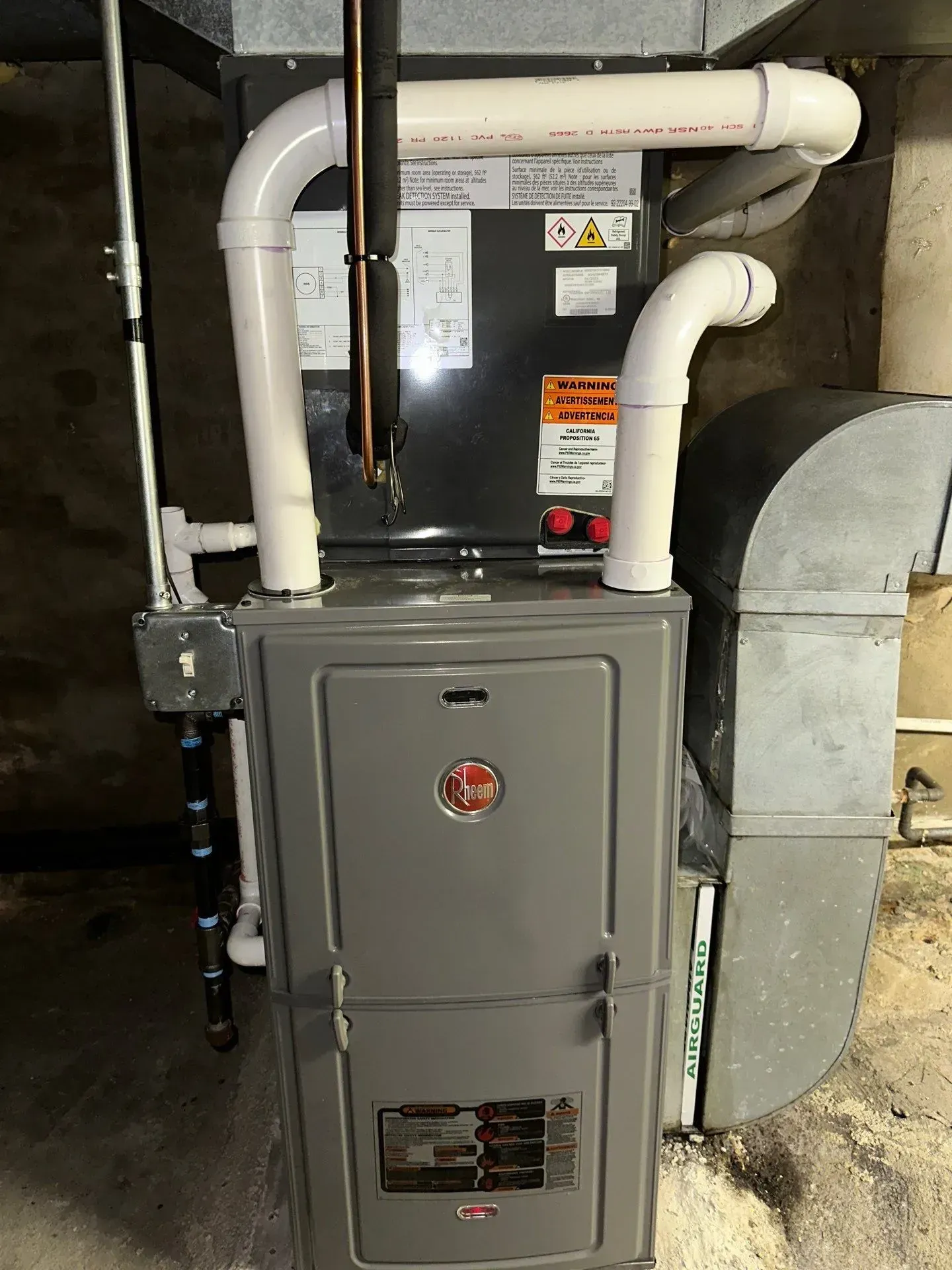 A gas furnace in a basement. The unit is gray with white PVC pipes extending from the top.