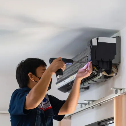 Man in mask repairs air conditioner with a drill, mounted on a white wall.