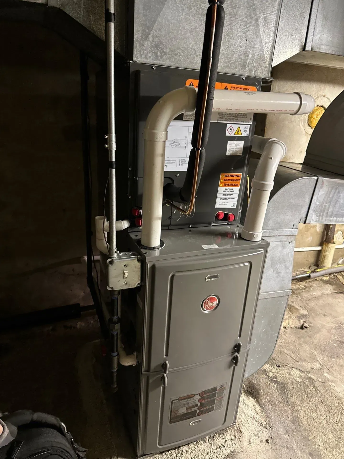 A gray Rheem furnace in a basement, with connected white pipes and ductwork.