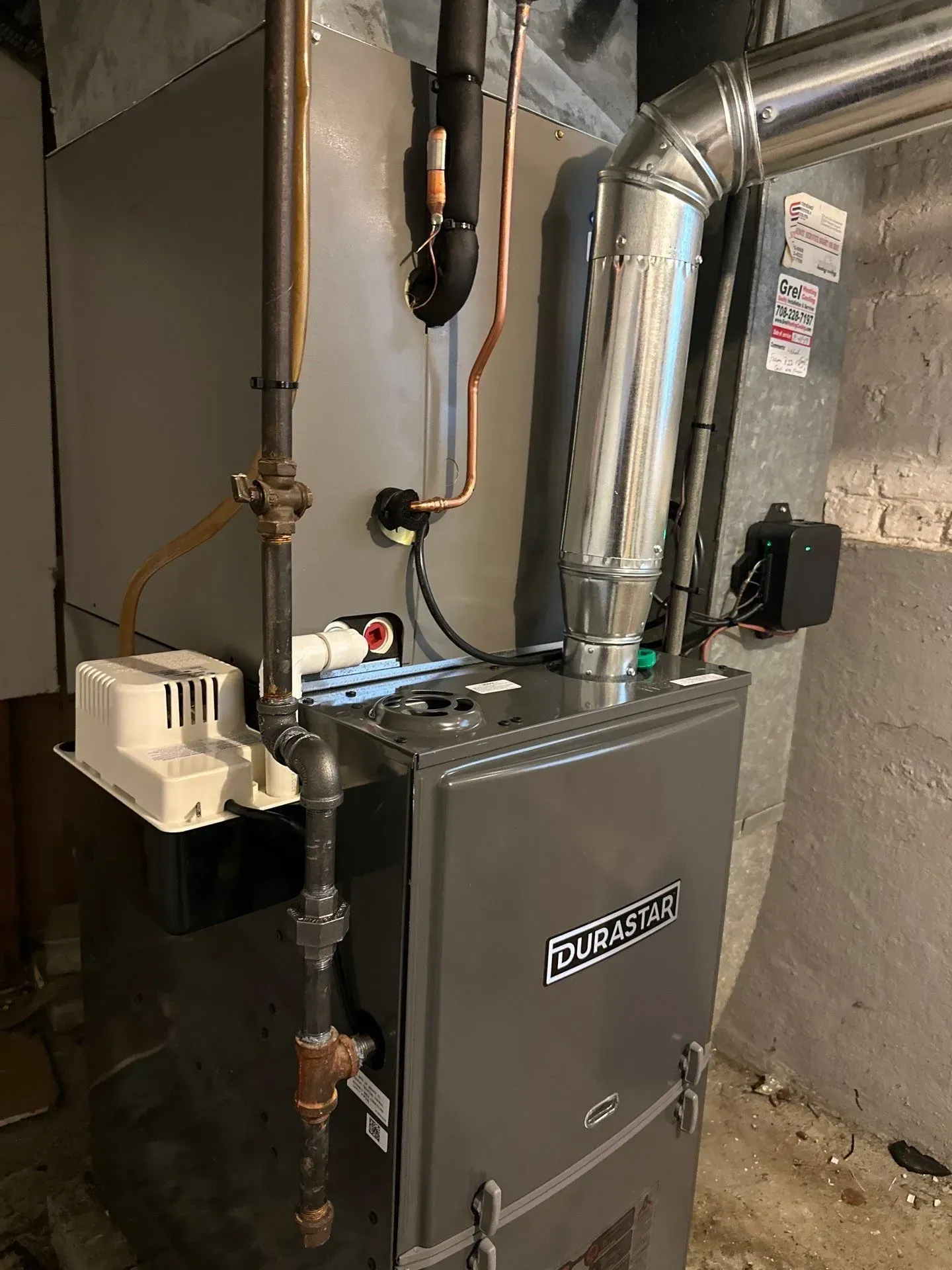 A gas furnace in a basement, with vent pipes and gas lines. Gray furnace, gray walls.