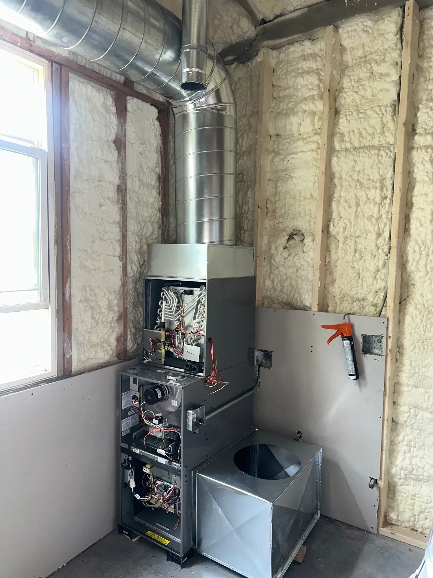 Furnace installed in a room with exposed wood framing and spray foam insulation; ductwork and vent pipe attached.