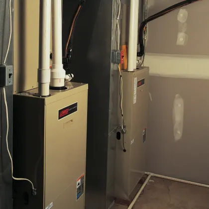 Two beige furnaces in a utility room. White pipes, black wires, and a gray wall.