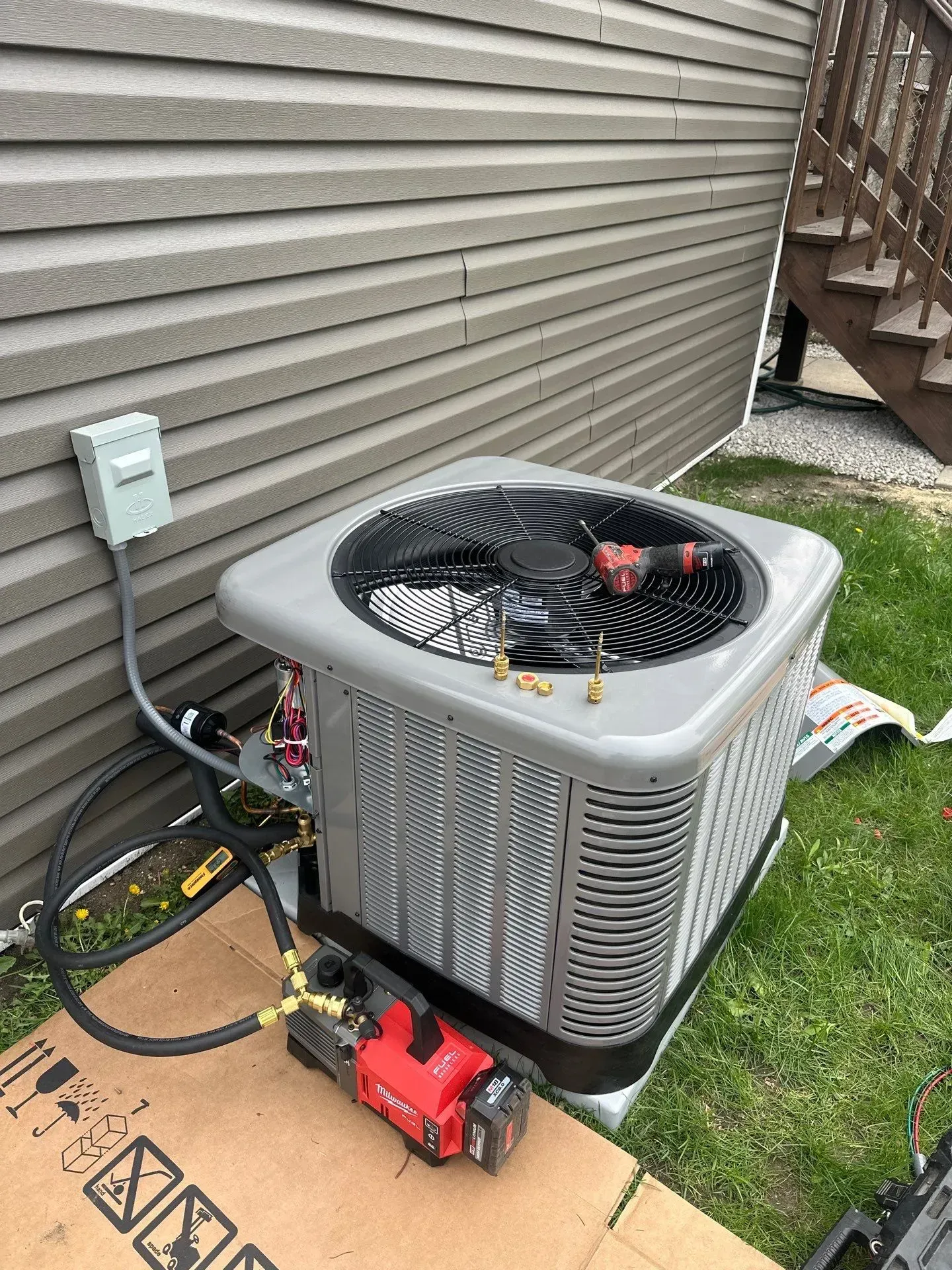 HVAC unit beside a house; a technician works on it with tools and hoses.