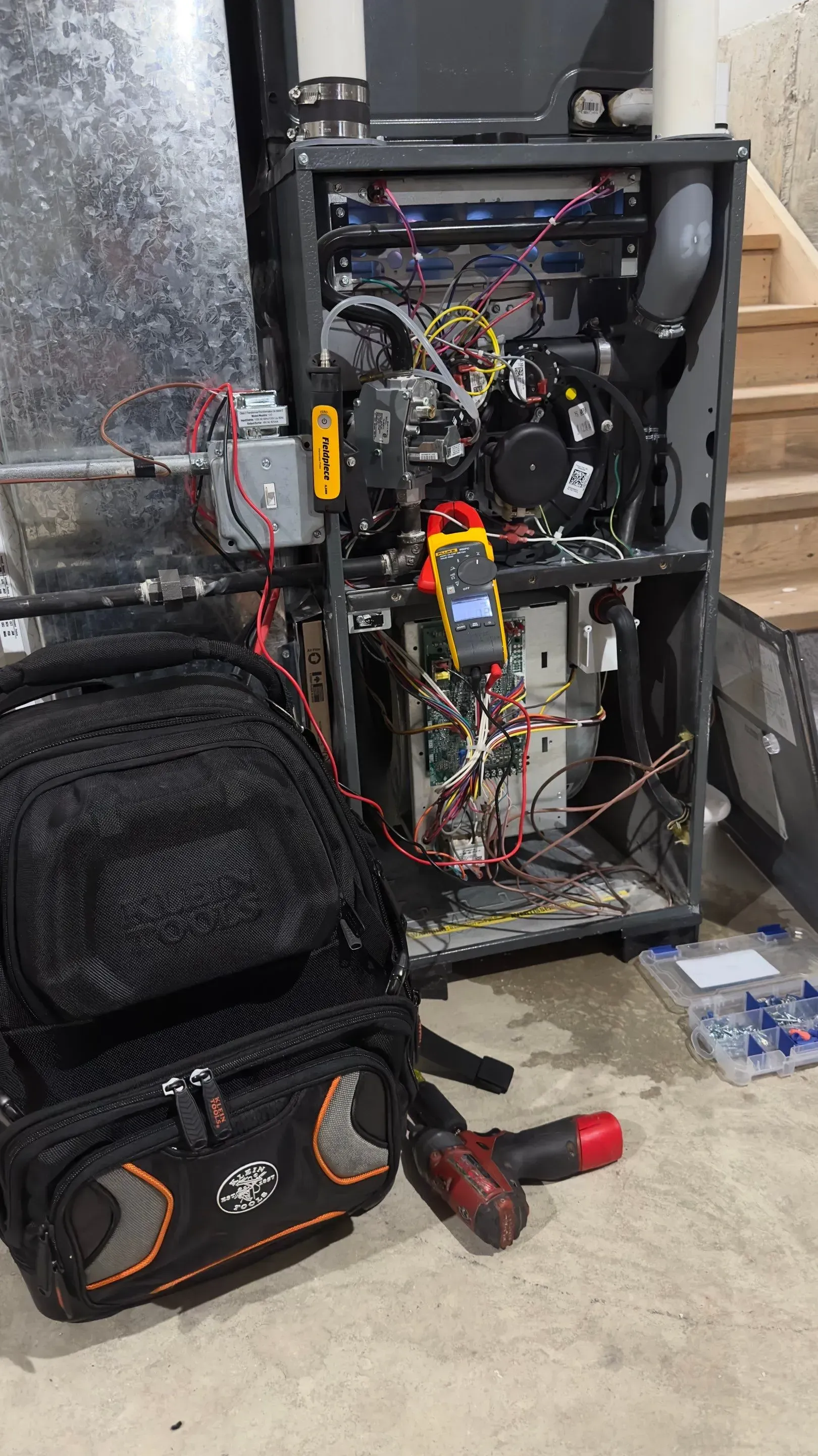 HVAC technician repairing a furnace. A multimeter is in use with a tool bag and toolbox nearby.