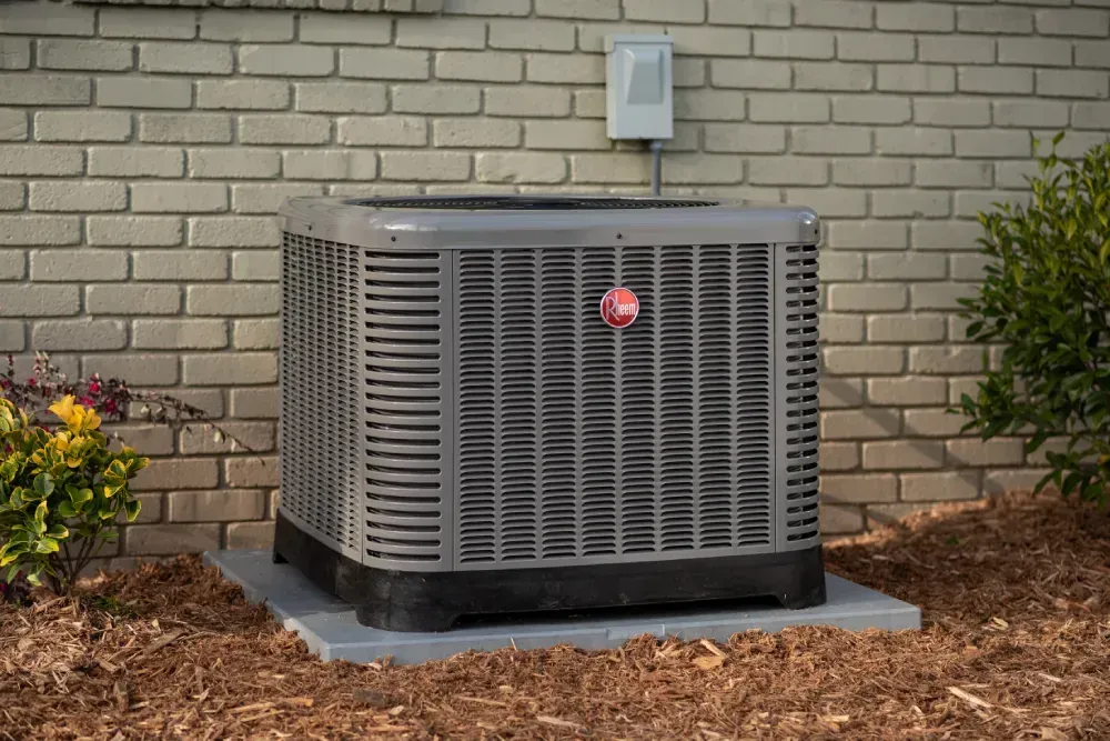 Gray Rheem air conditioning unit against a brick wall, on a concrete pad in a yard.