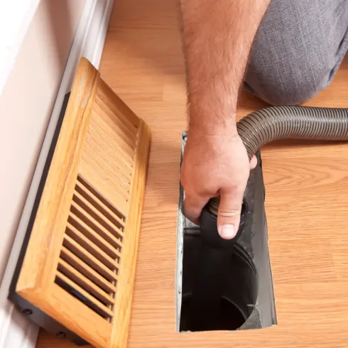 Person vacuuming a floor vent with a vacuum hose, wooden vent cover is off.