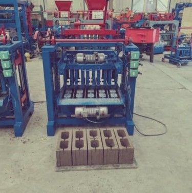 Blue brick-making machine in a workshop, with stacked brown concrete blocks in front.