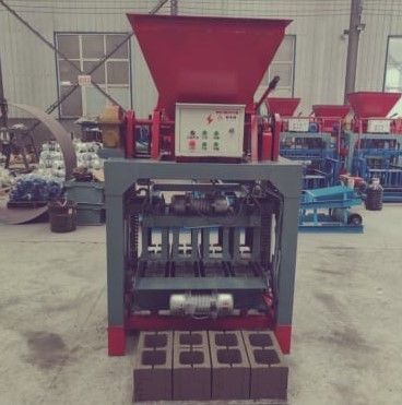Red industrial brick-making machine in a factory, with a hopper and fresh bricks on the floor.