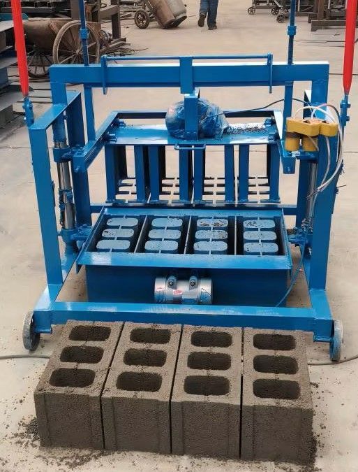 Blue brick-making machine pressing concrete blocks in a workshop, with finished blocks stacked in front.