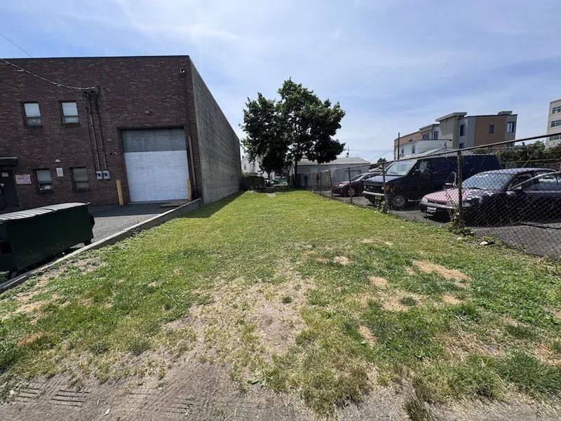 Empty grassy lot beside brick building and parked cars under a partly cloudy sky