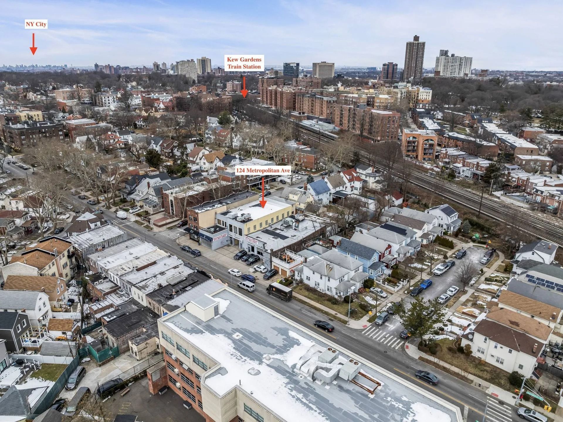 Aerial view of a snowy neighborhood with houses, streets, and distant apartment buildings under a clear sky.