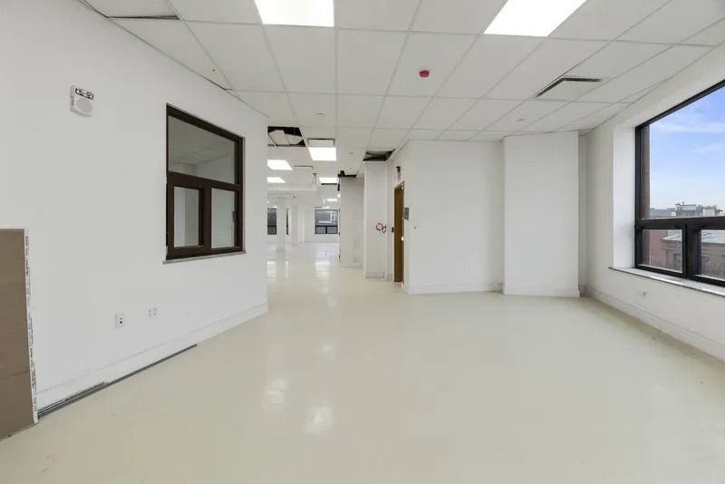 Bright empty office hallway with white floors, ceiling lights, and large windows on the right.
