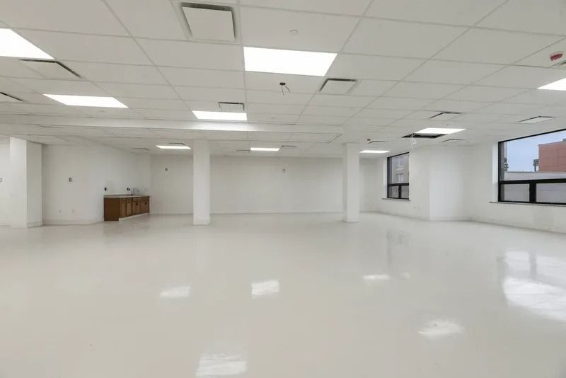 Empty white office room with glossy floors, drop ceiling lights, and windows along the right wall