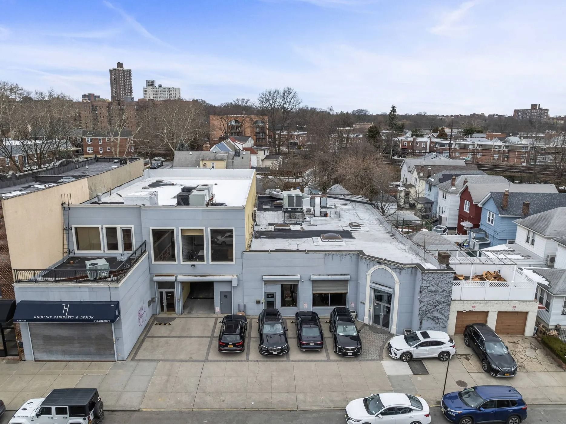Aerial view of a snowy urban block with rooftops, parked cars, and low-rise buildings.