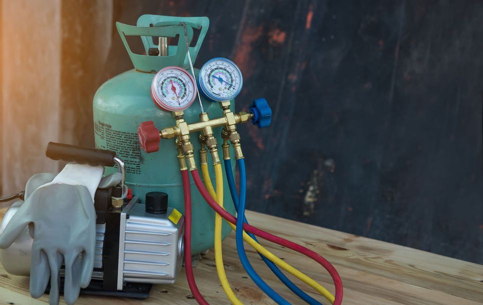 Refrigeration equipment: gauge manifold, tank, vacuum pump, hoses, gloves on wood surface.