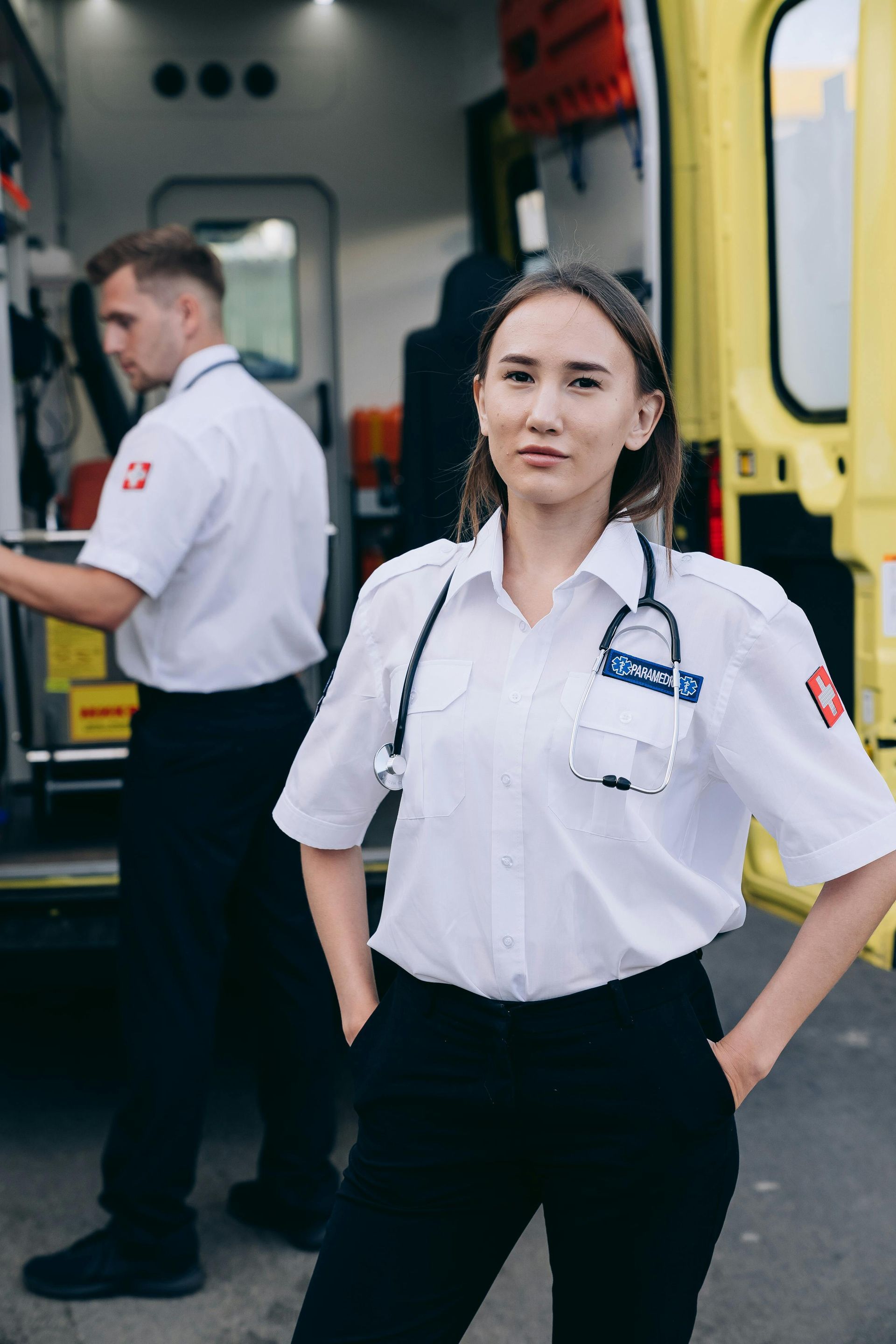 Paramedic stands confidently in front of an ambulance, hands in pockets. Another medic in the background.