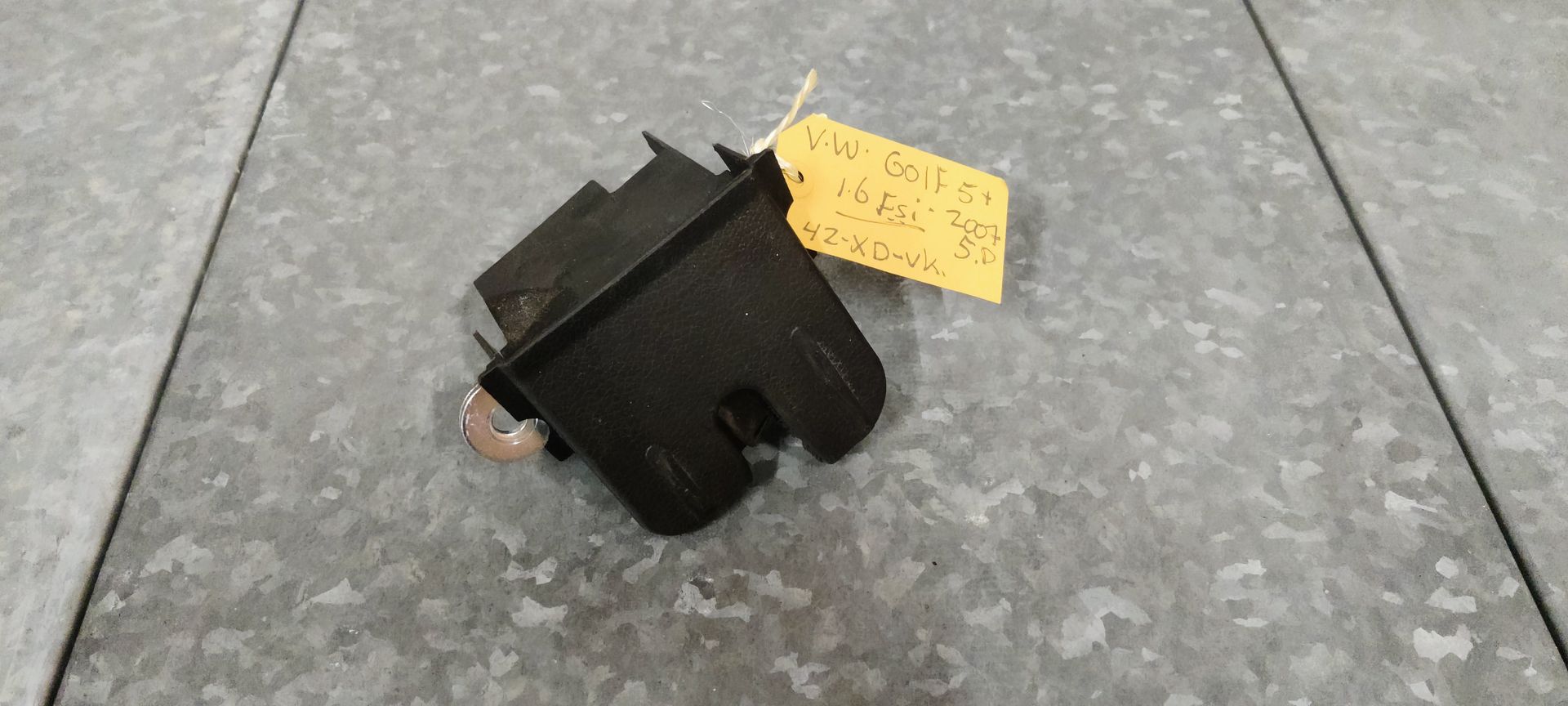 Een zwart kofferbakslot van een auto ligt op een grijze tegelvloer, vergezeld van een klein geel identificatielabeltje.