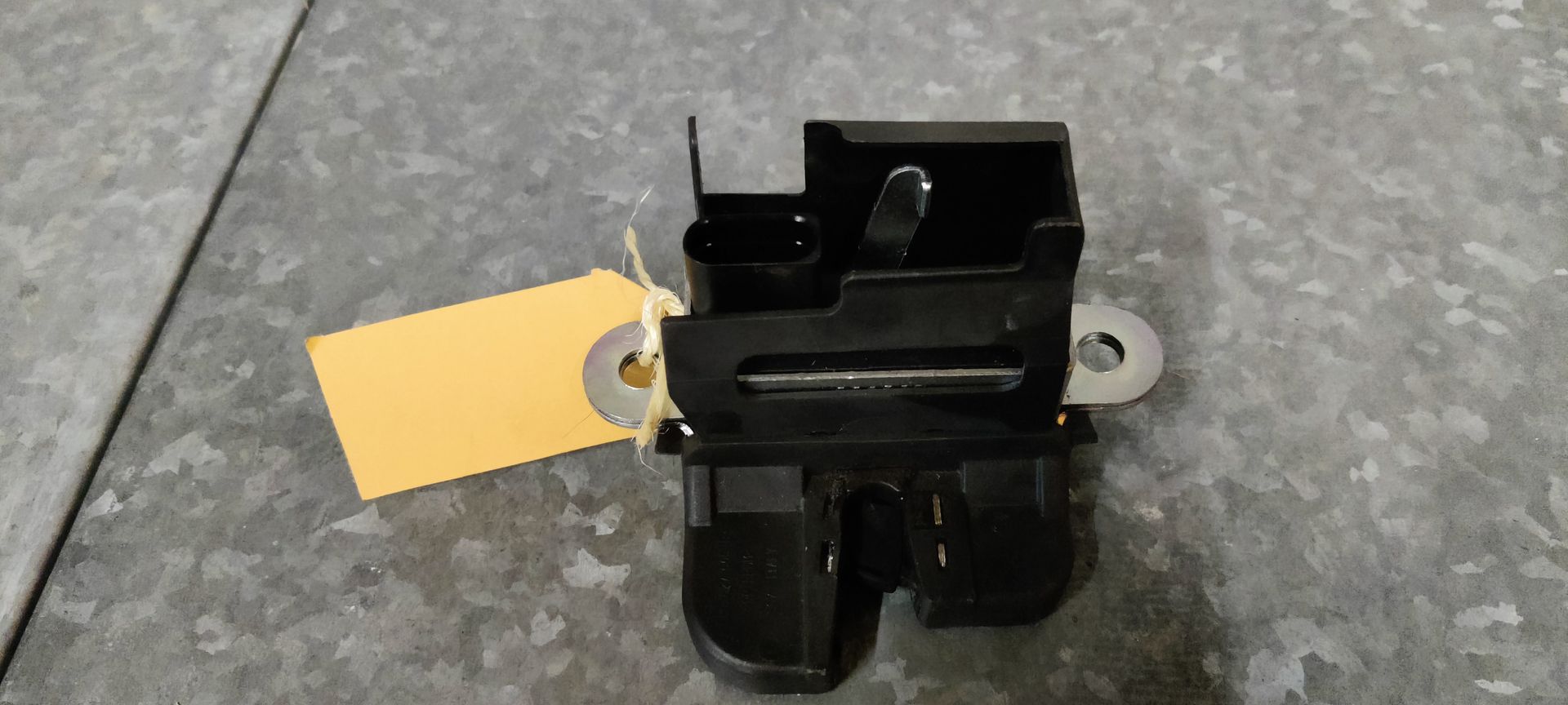 Een zwart kofferbakslot van een auto ligt op een grijze tegelvloer, vergezeld van een klein geel identificatielabeltje.