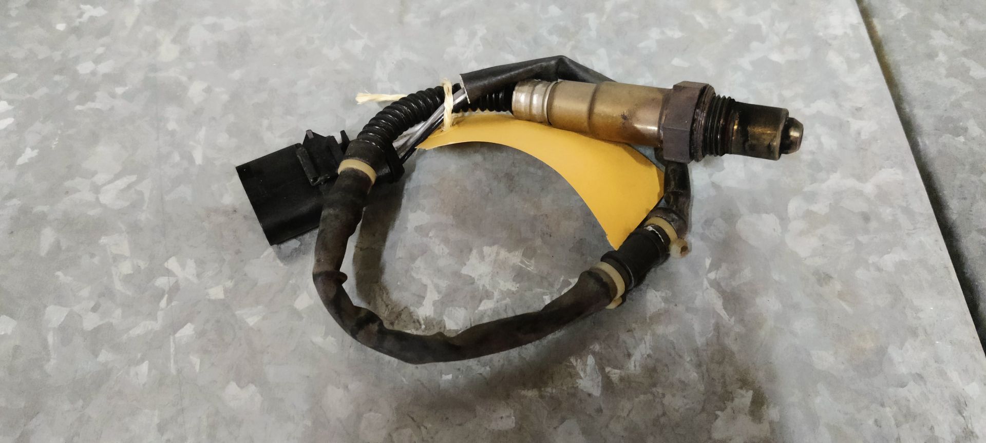 Een zuurstofsensor voor auto's met een zwarte kabelboom en connector, liggend op een lichtgekleurde ondergrond met een geel label.