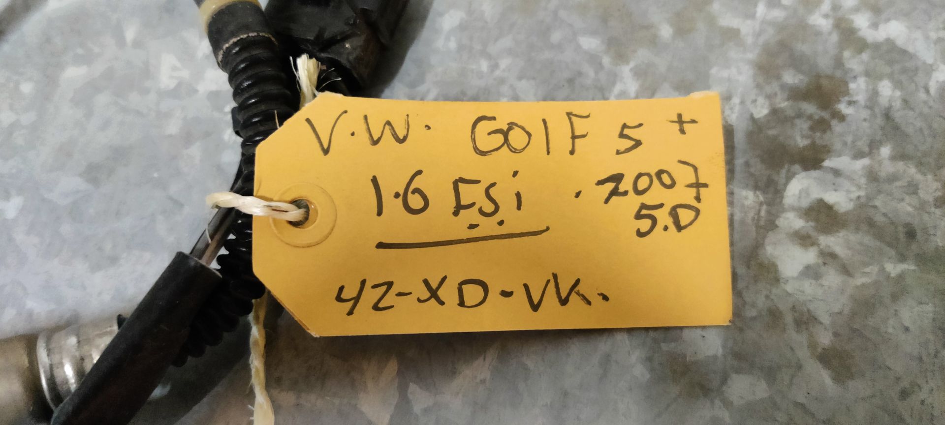 Een zuurstofsensor voor auto's met een zwarte kabelboom en connector, liggend op een lichtgekleurde ondergrond met een geel label.