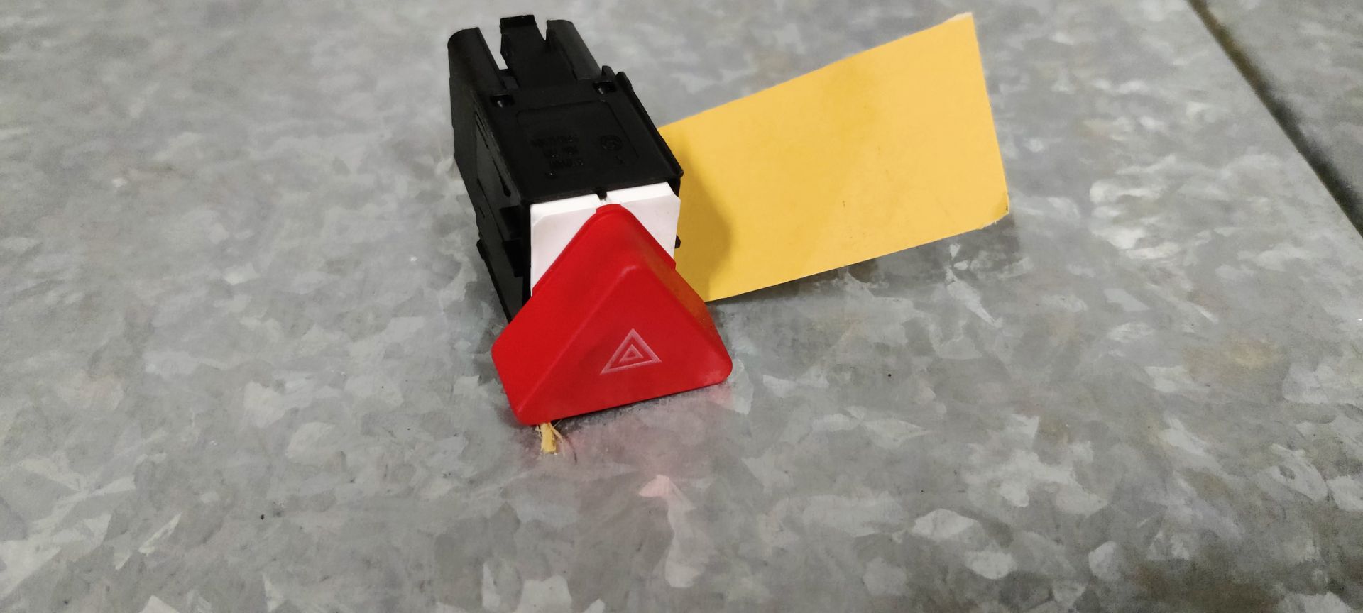 Een rode driehoekige waarschuwingslichtschakelaar met een wit pictogram en een geel label, geplaatst op een metalen oppervlak.