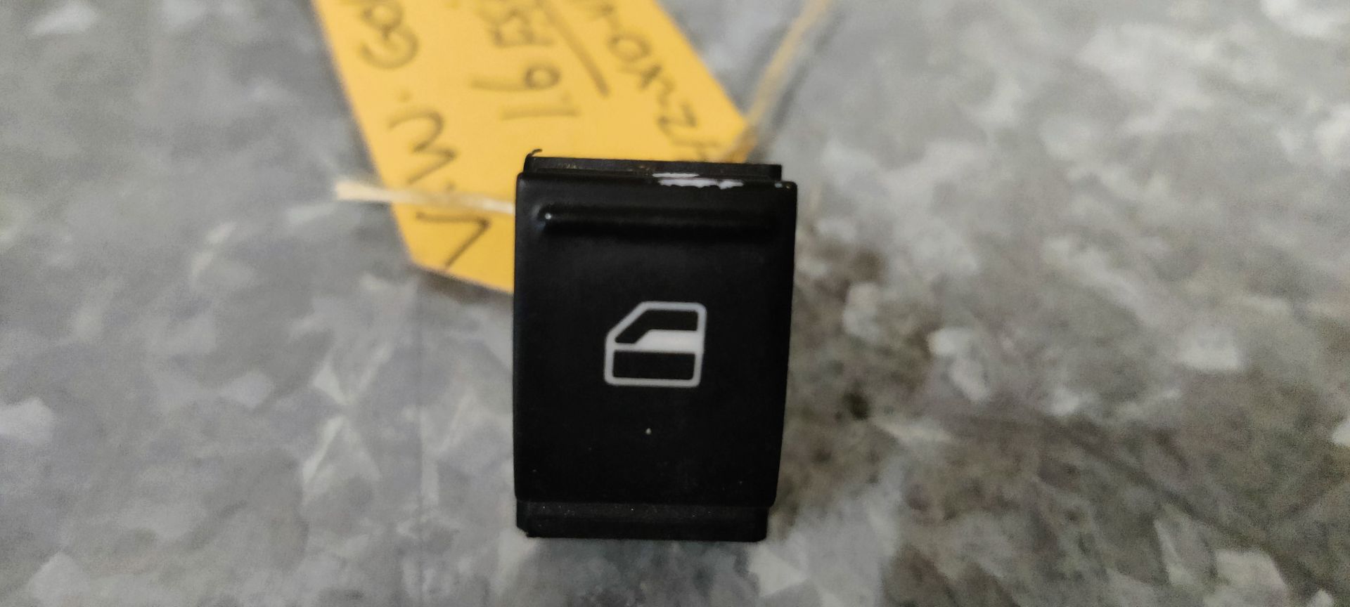 Een enkele zwarte Volkswagen-schakelaar voor de elektrische ramen, voorzien van een wit raamicoon, rustend op een grijs, gestructureerd oppervlak.