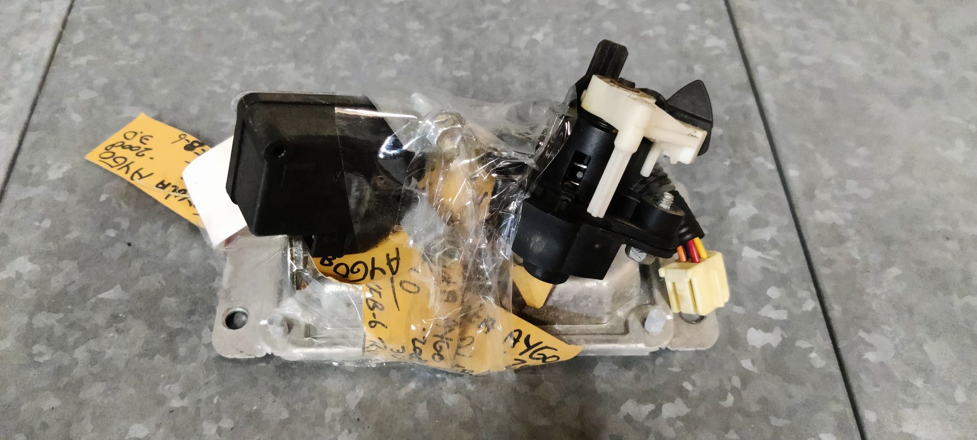 Een actuatoreenheid voor het vergrendelen van een autodeur, met een kunststof behuizing en draadconnectoren, gemonteerd op een kleine metalen beugel.