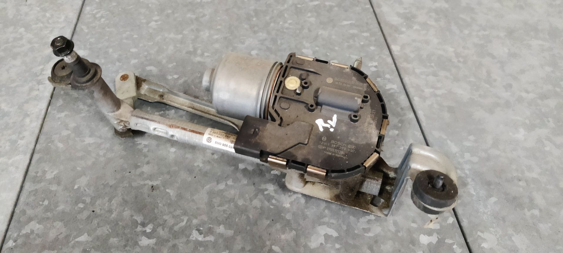 Een ruitenwissermotor met metalen arm en zwarte motorbehuizing, rustend op een grijze tegelondergrond.