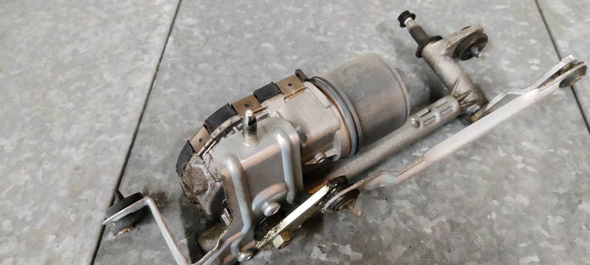 Een ruitenwissermotor met metalen arm en zwarte motorbehuizing, rustend op een grijze tegelondergrond.