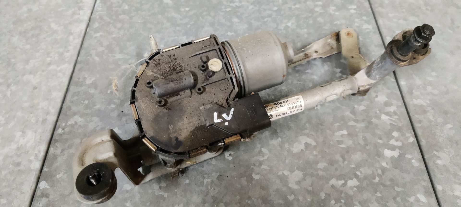 Een gebruikte ruitenwissermotor met bijbehorende koppeling op een grijze werkplaatsvloer.