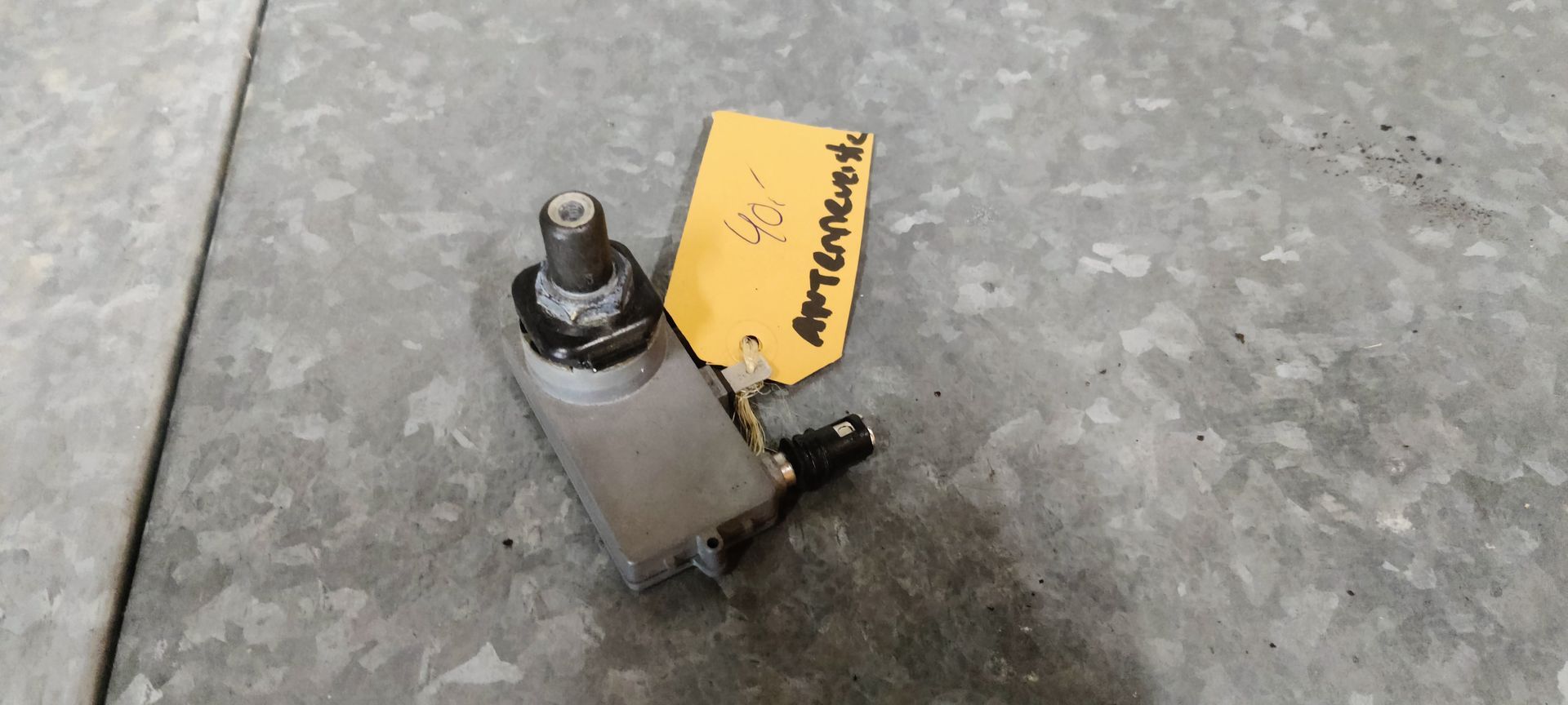 Een kleine, metalen autosensor met een schroefdraad en een geel labeltje op een grijs, gestructureerd oppervlak.
