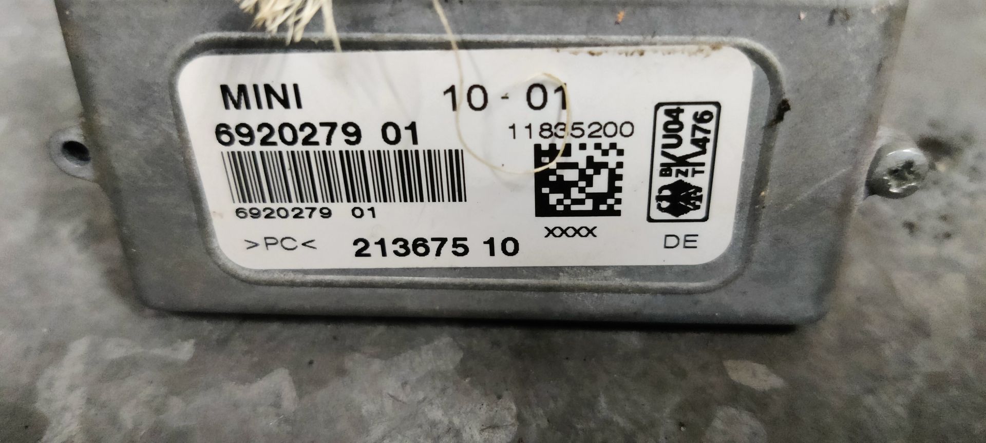 Een kleine, metalen autosensor met een schroefdraad en een geel labeltje op een grijs, gestructureerd oppervlak.