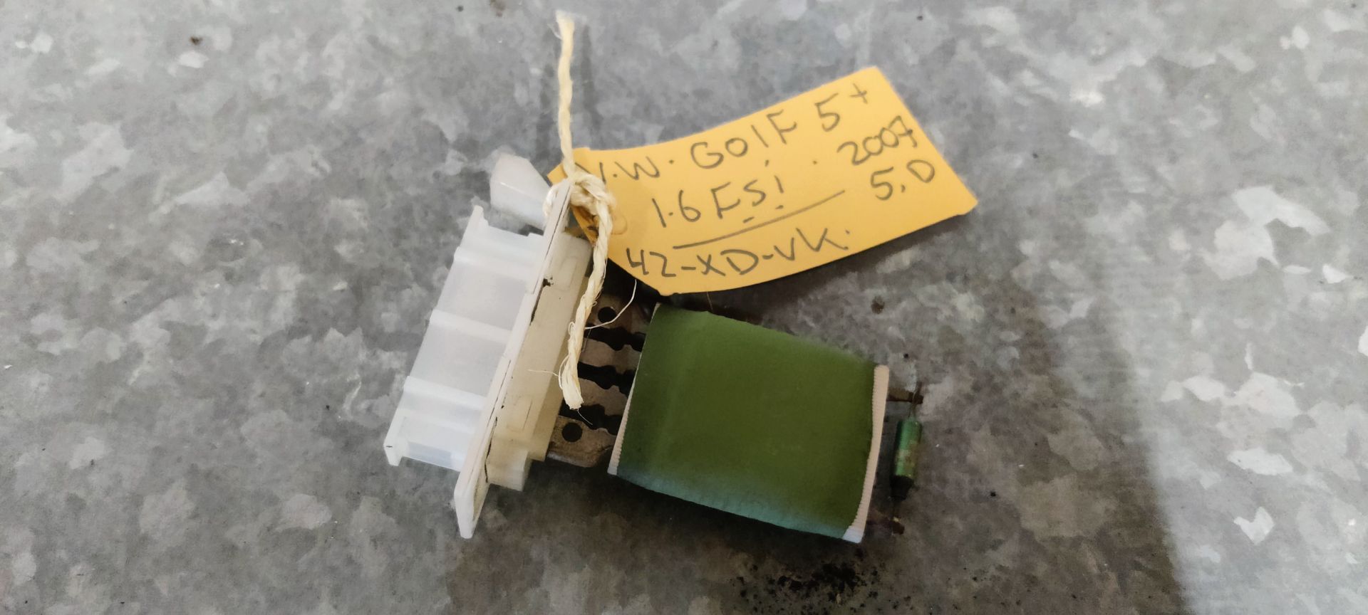 Een groene weerstand voor de ventilator van de airconditioning van een Volkswagen Golf 5 uit 2007, met een witte plastic connector op een metalen oppervlak.