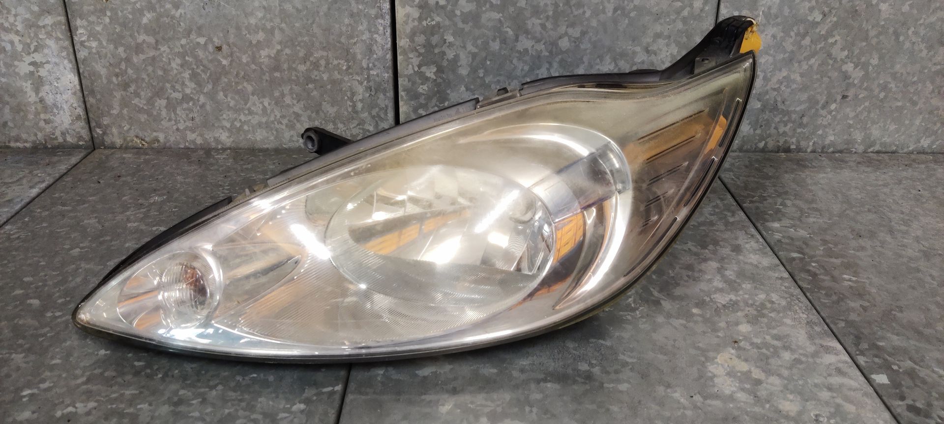 Een gebruikte, gebogen autokoplamp met een heldere lens en interne reflectoren, geplaatst tegen een grijze tegelachtergrond.