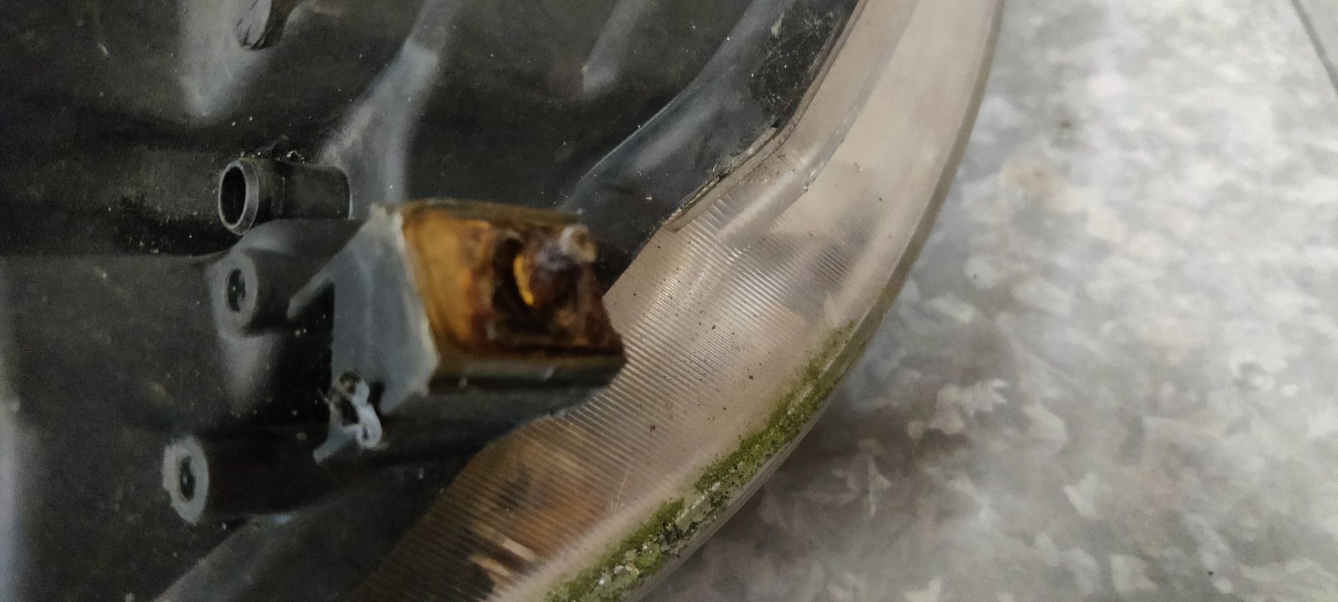 Een gebruikte, gebogen autokoplamp met een heldere lens en interne reflectoren, geplaatst tegen een grijze tegelachtergrond.