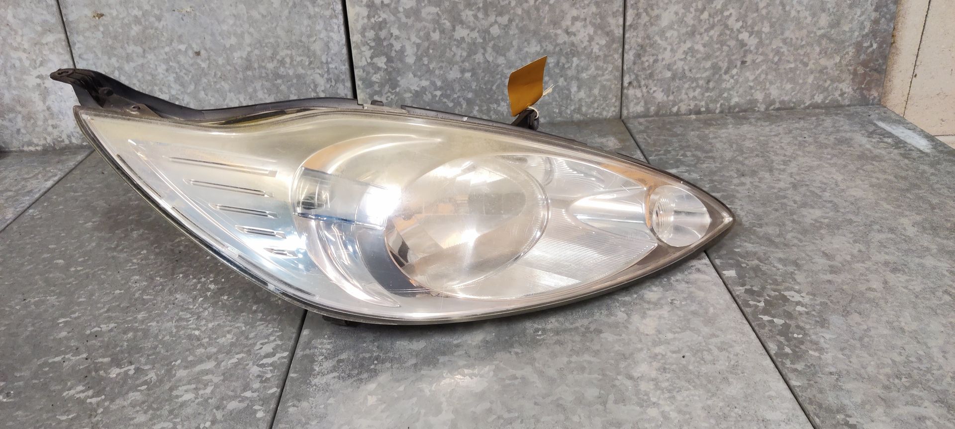 Een gebogen autokoplampunit met een heldere lens en interne reflector, geplaatst op een grijze tegelondergrond.