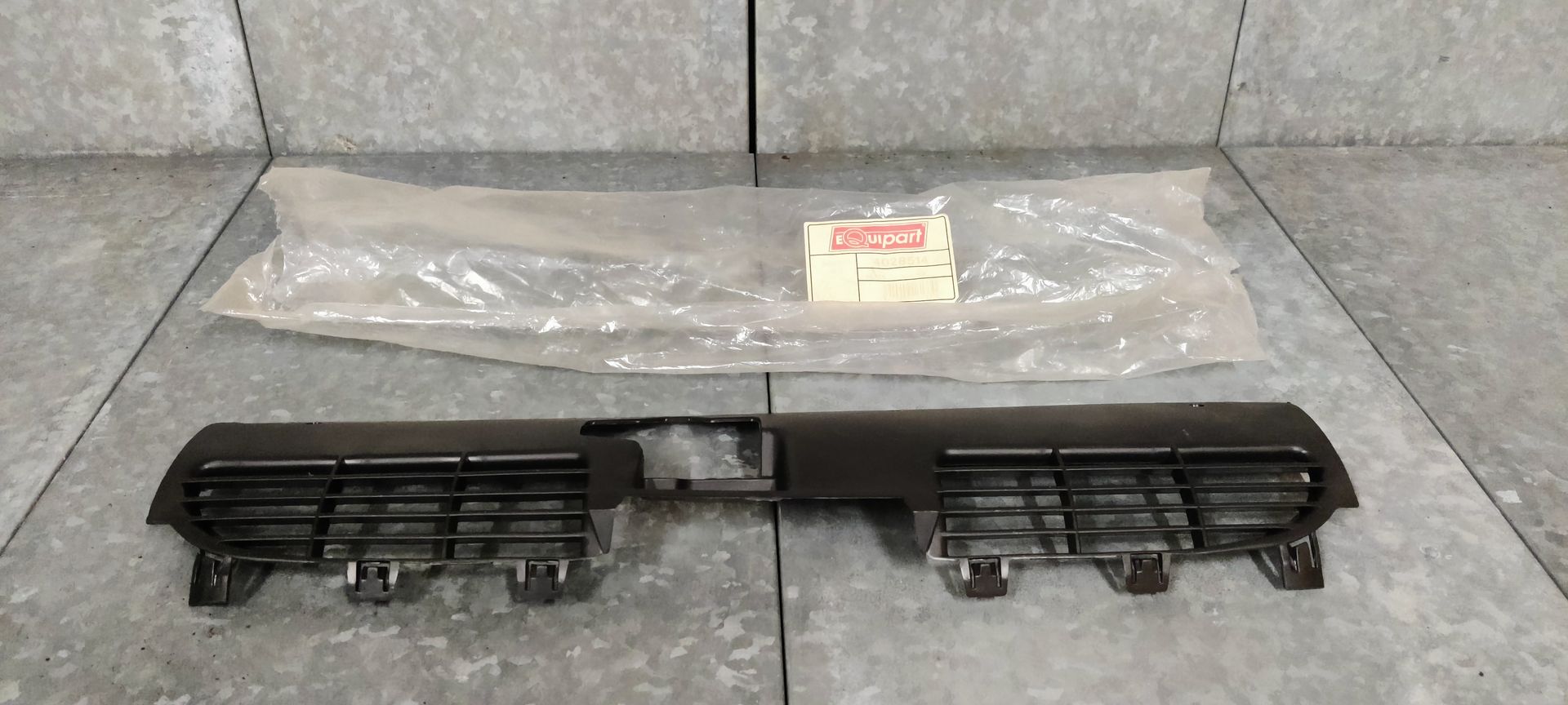 Een matzwarte afdekking voor een autoradiatorrooster met uitsparingen in het midden, geplaatst tegen een grijze achtergrond.