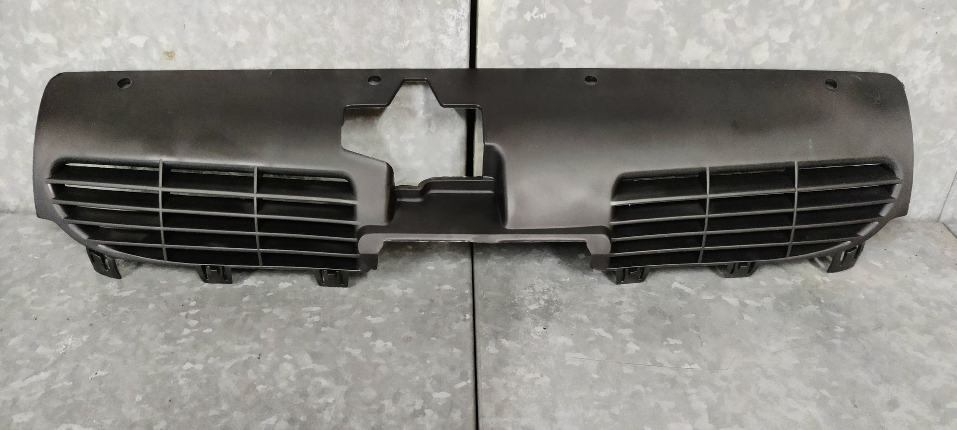 Een matzwarte afdekking voor een autoradiatorrooster met uitsparingen in het midden, geplaatst tegen een grijze achtergrond.