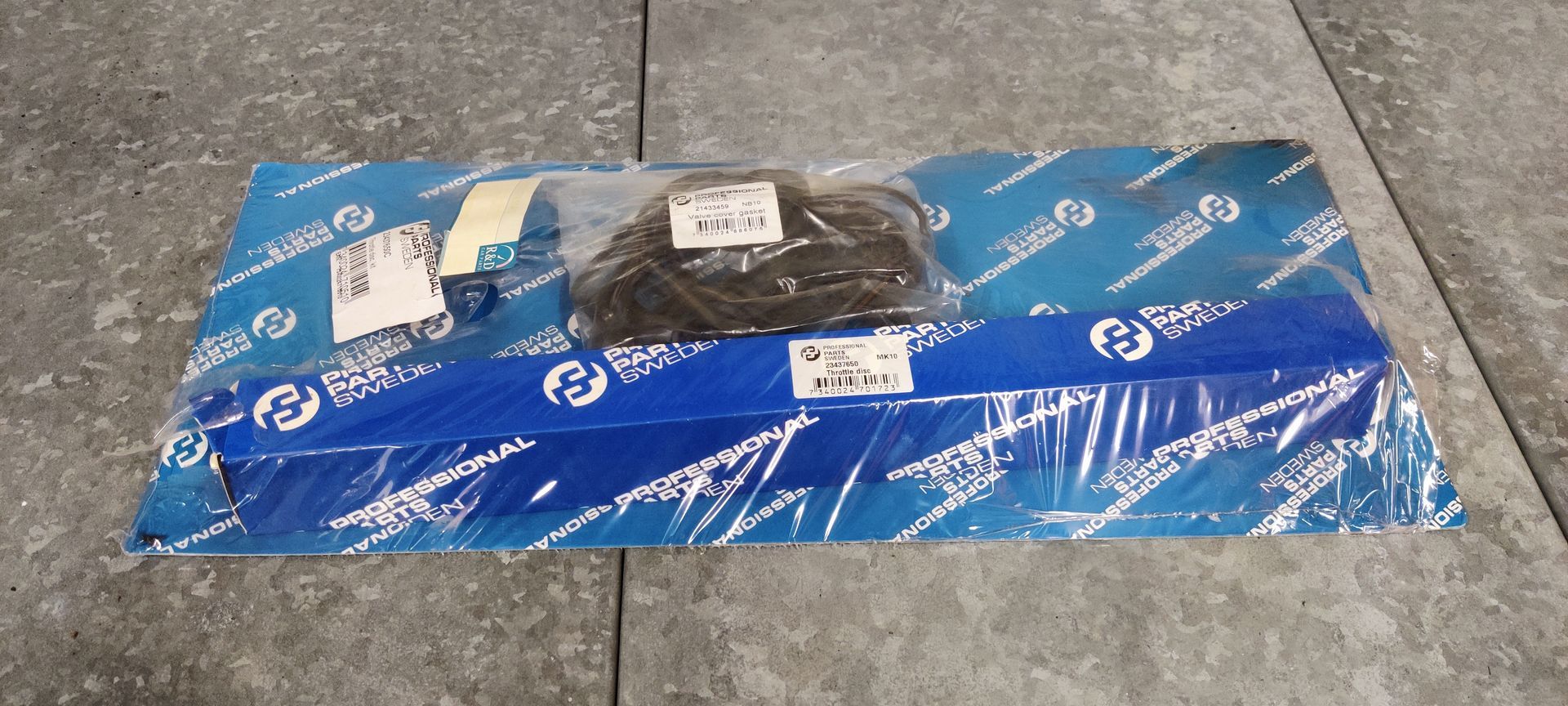 Een verzegelde blauw-witte verpakking met een distributiekettingrevisieset voor auto's, op een grijze betonnen ondergrond.