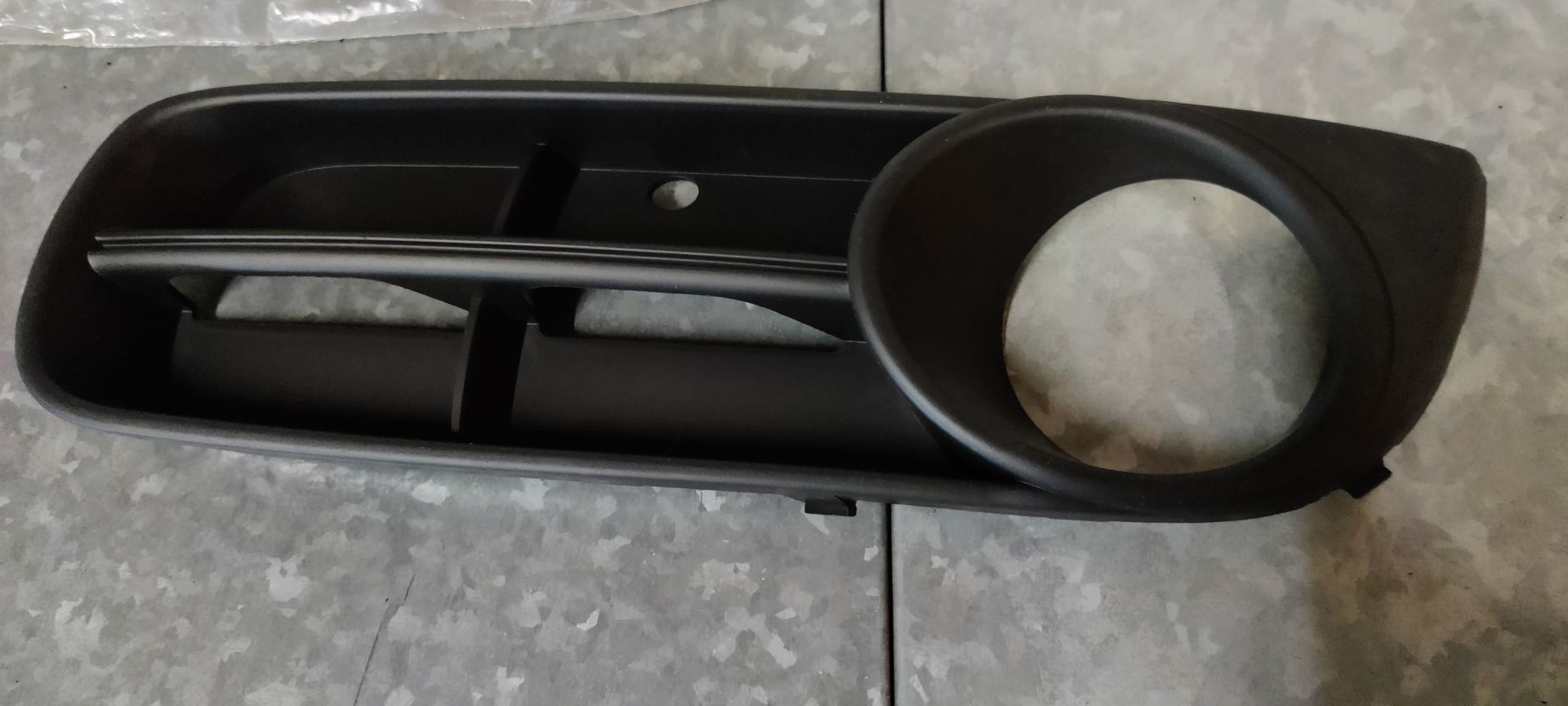 Zwarte kunststof bumper met mistlampgrille, ronde lampbehuizing en horizontale lamellen, geplaatst tegen een betonnen vloer.