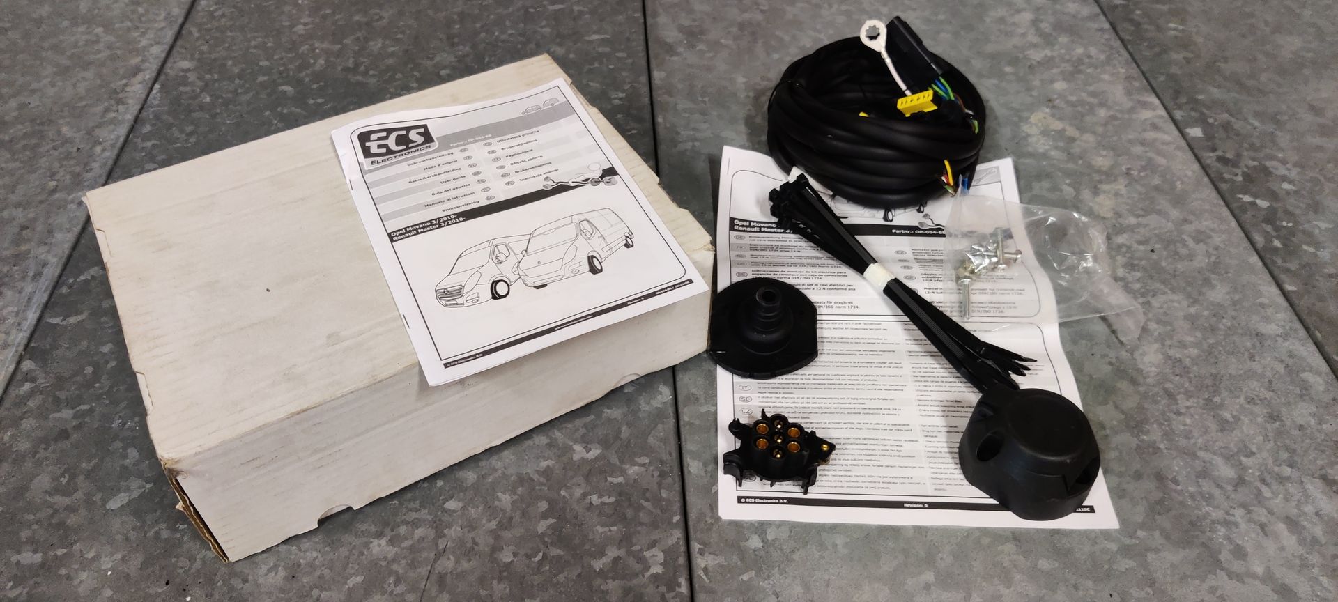 Opel Movano trekhaak. Een in een doos verpakte kabelboomset voor een trekhaak, inclusief instructies, elektrische kabels, een stekker en klein bevestigingsmateriaal, ligt op een tegelvloer.