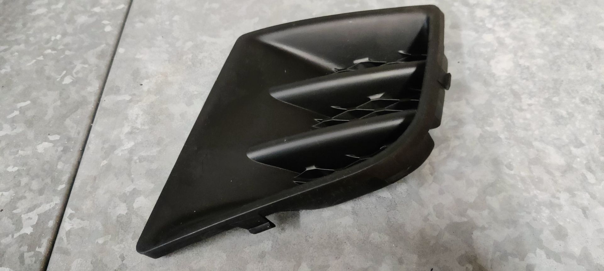 Een zwart plastic inzetstuk voor een mistlampgrille, met een driehoekige vorm en horizontale lamellen.