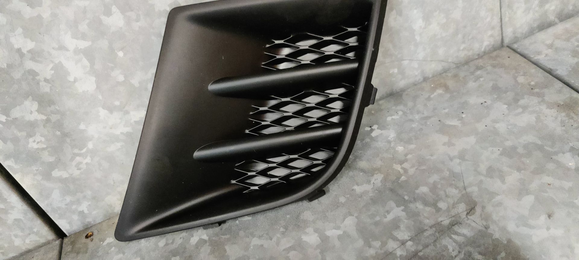 Een zwart plastic inzetstuk voor een mistlampgrille, met een driehoekige vorm en horizontale lamellen.